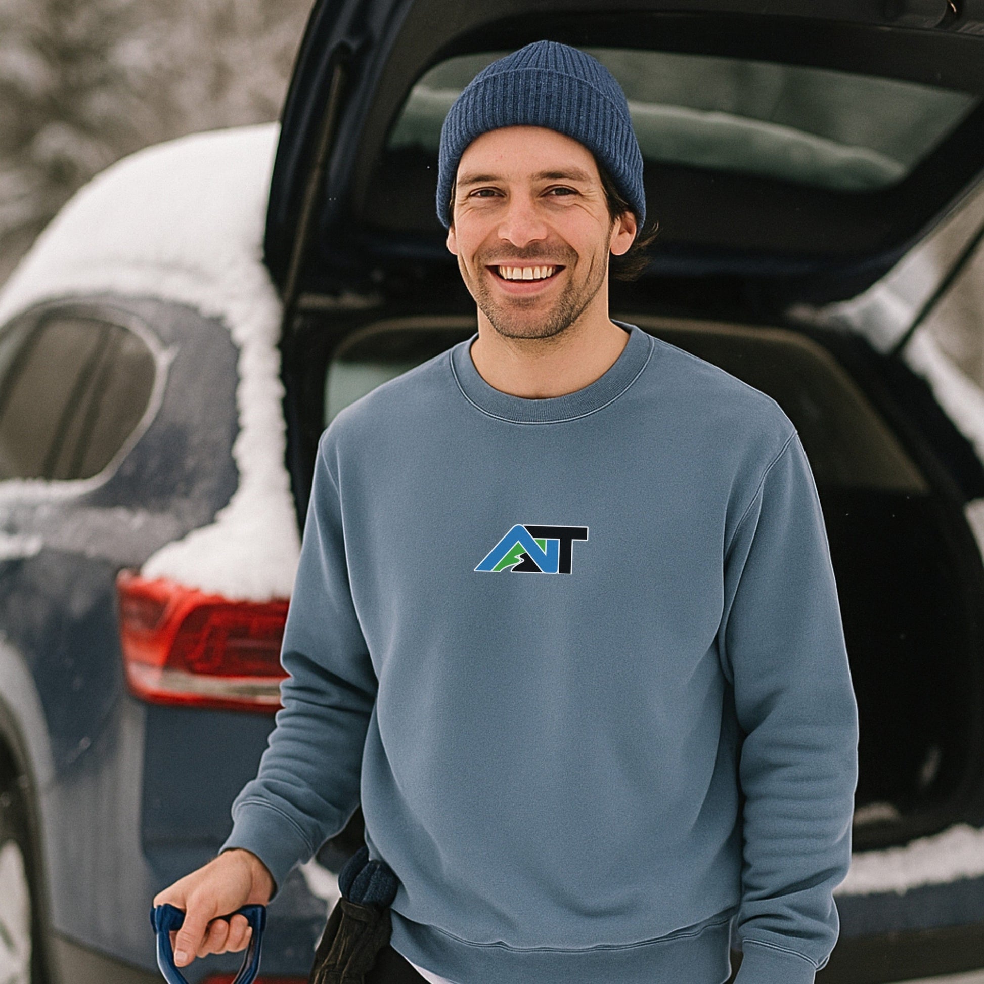 Smiling man in blue beanie and cool blue AT sweatshirt standing next to snow-covered car, holding ski gear in winter parking lot