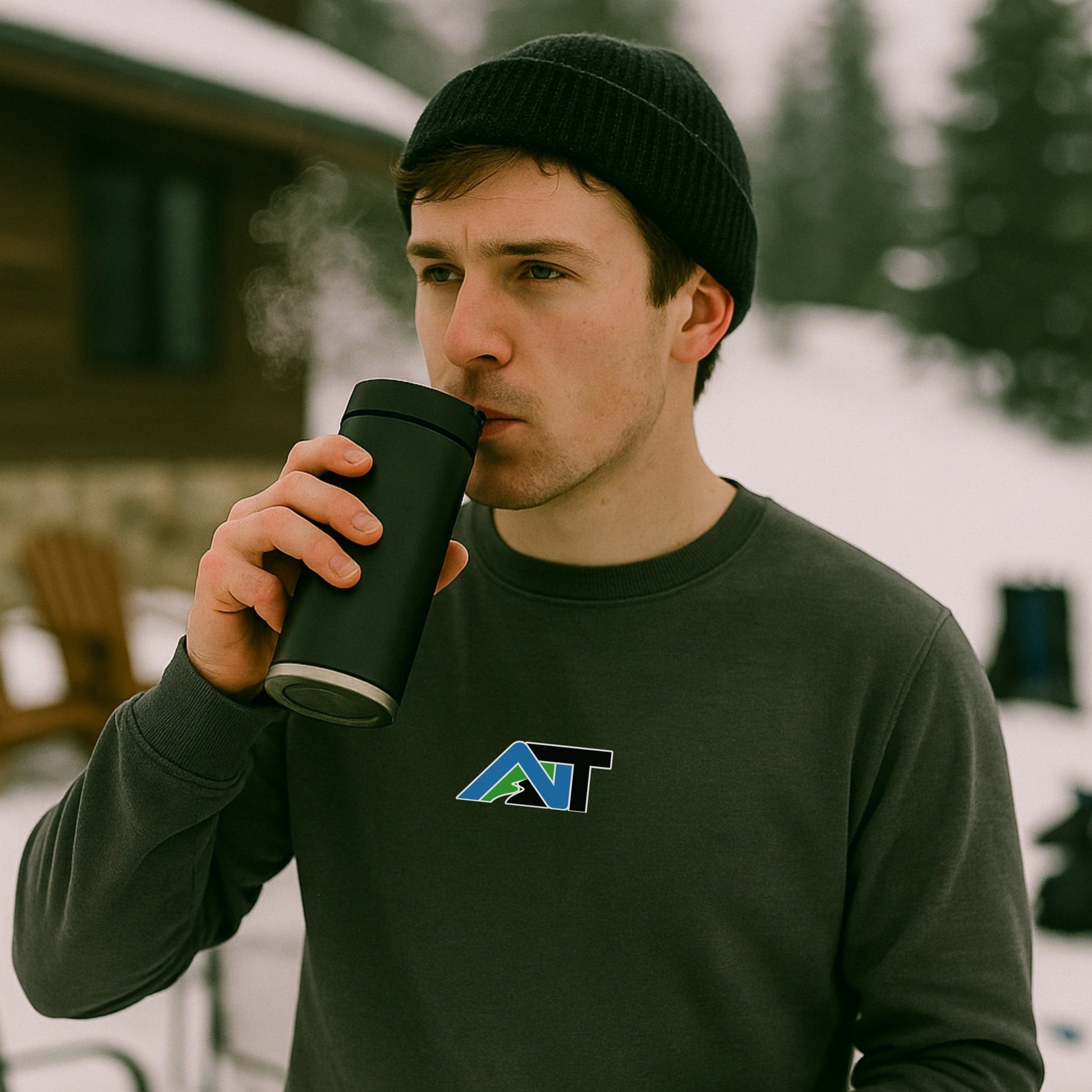 Man in dark beanie and pepper-colored AT sweatshirt sipping hot drink from black travel mug outside snowy ski cabin