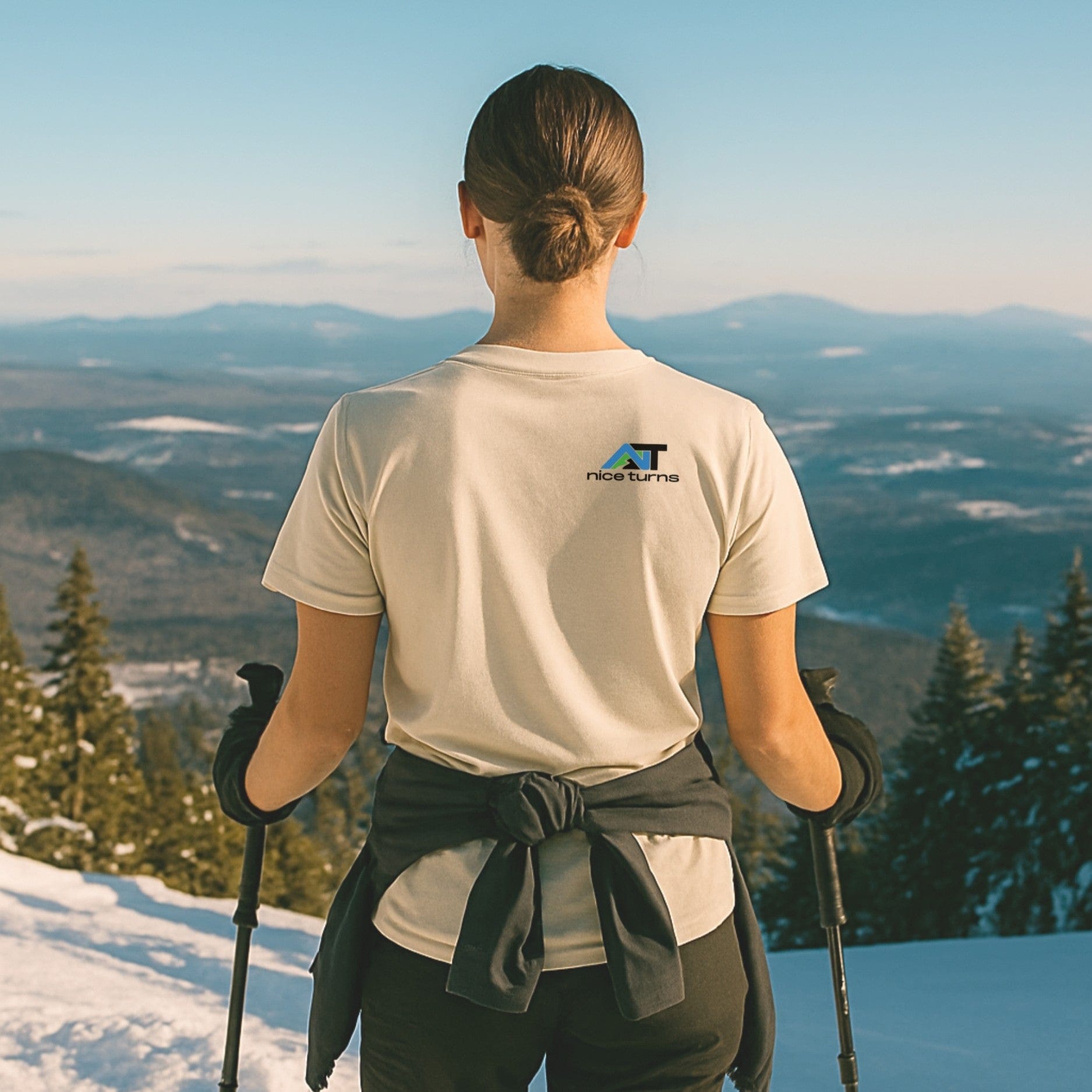 Cream T-shirt with blue and black “nice turns” logo on back, worn by skier with poles on a snowy ridge overlooking distant mountains.