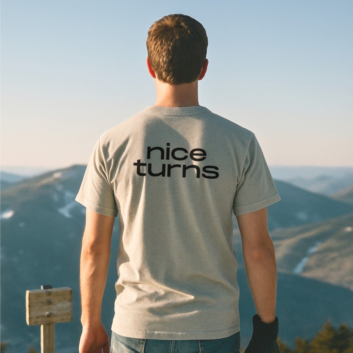 Light grey nice turns T-shirt with bold back logo, worn by skier overlooking distant snowy mountain ridges at golden hour.