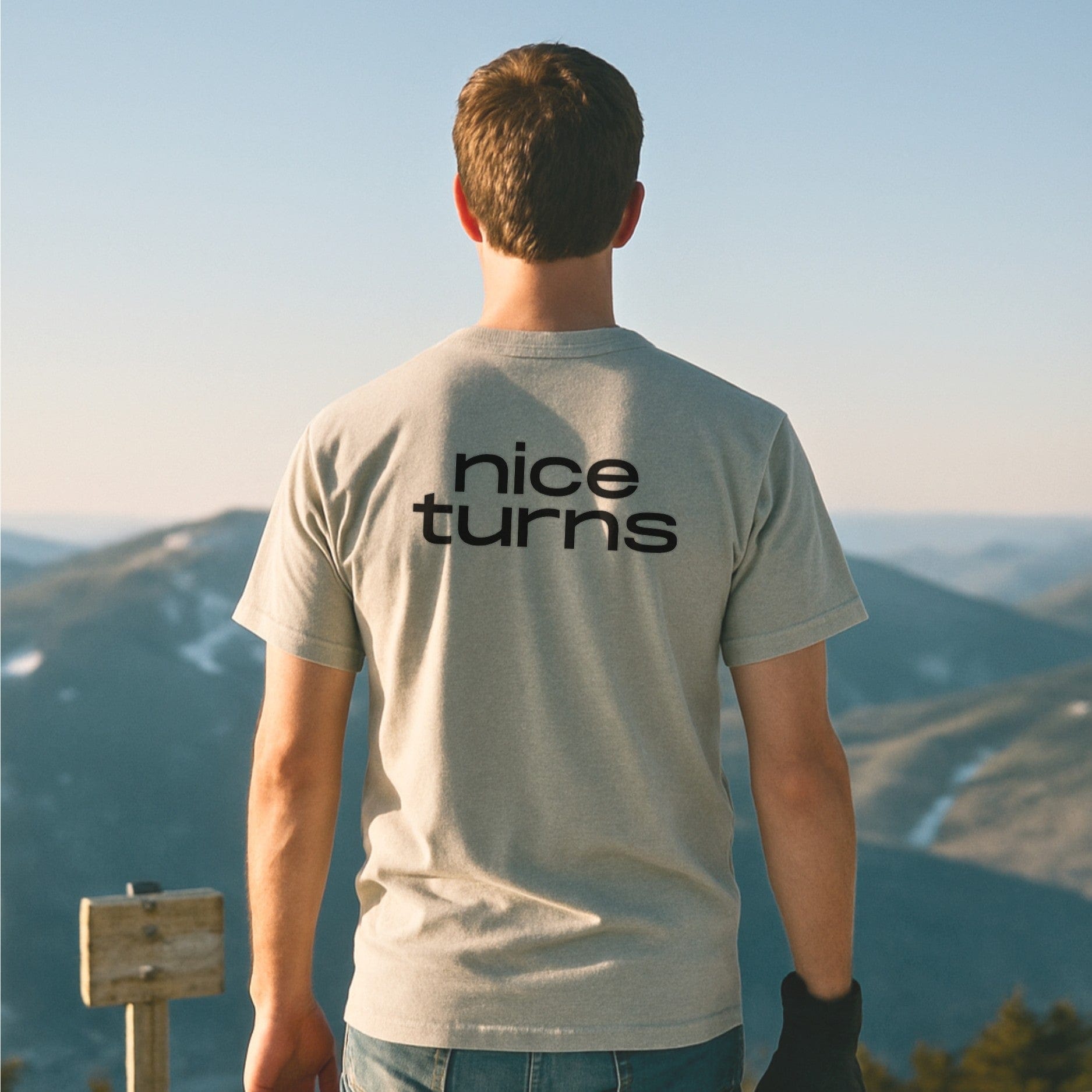 Light grey nice turns T-shirt with bold back logo, worn by skier overlooking distant snowy mountain ridges at golden hour.