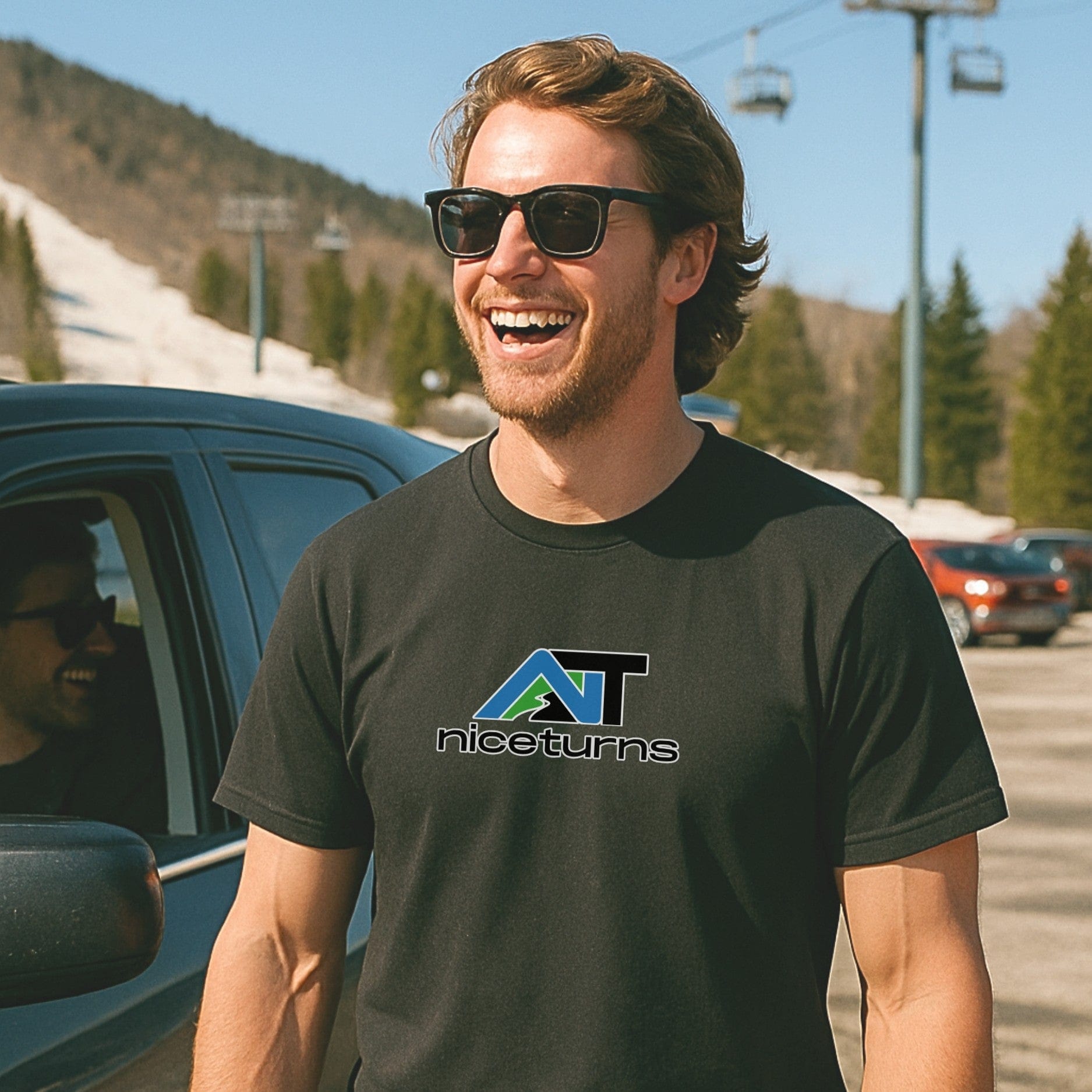 Black t-shirt featuring the alpine-themed nice turns logo, worn in a sunny ski resort parking lot with spring snow still on the slopes.