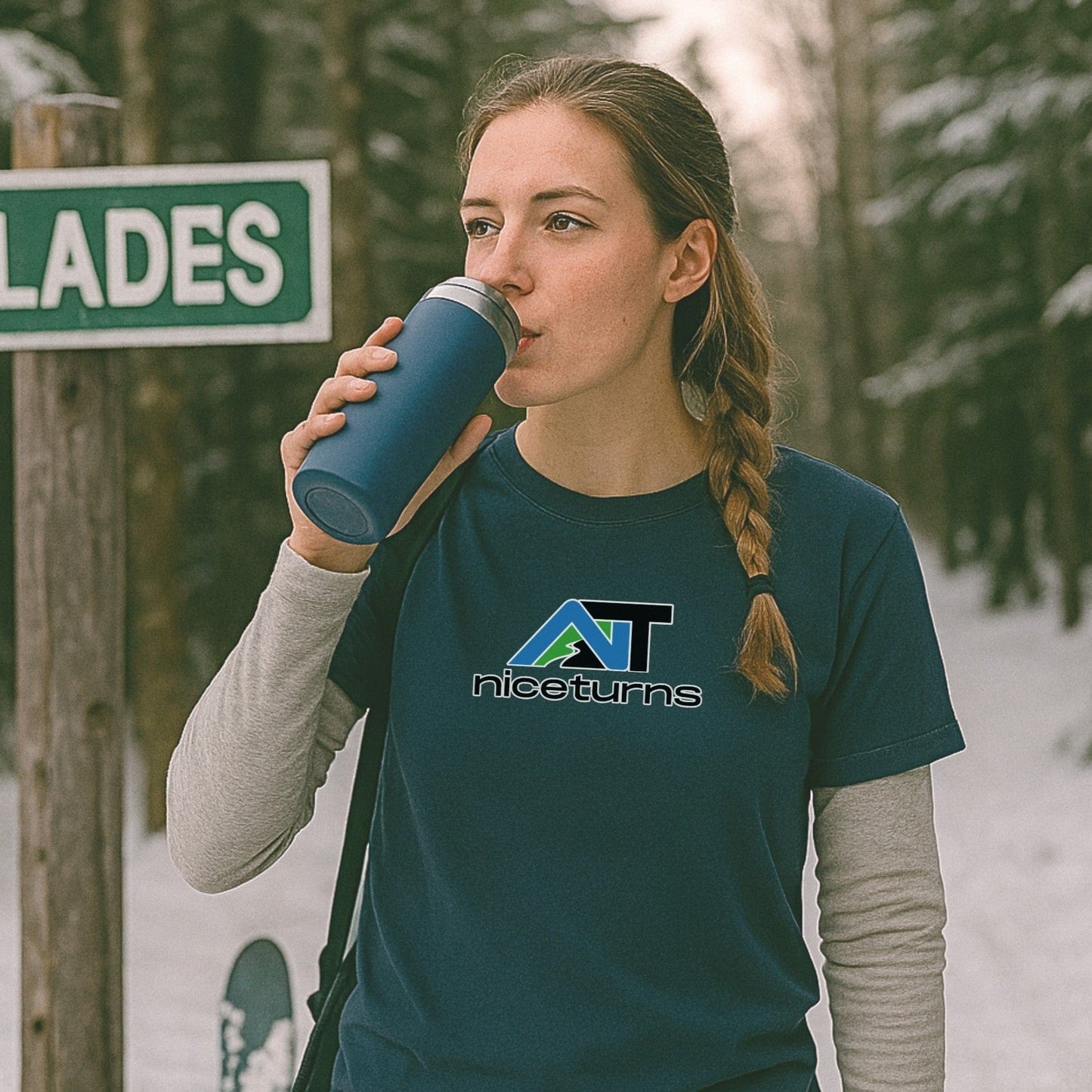 Indigo nice turns alpine logo t-shirt layered over a long sleeve, worn trailside near glades sign with a thermos and skis in the snow.