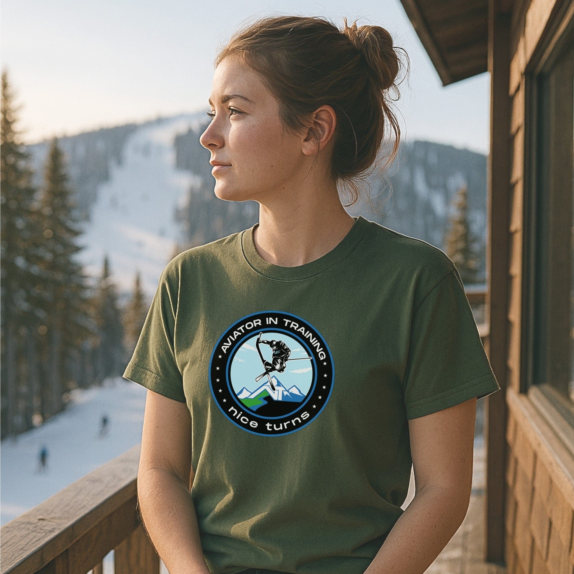 Aviator in Training tee from nice turns in military green, worn on a ski lodge deck overlooking a snowy mountain trail at sunset.