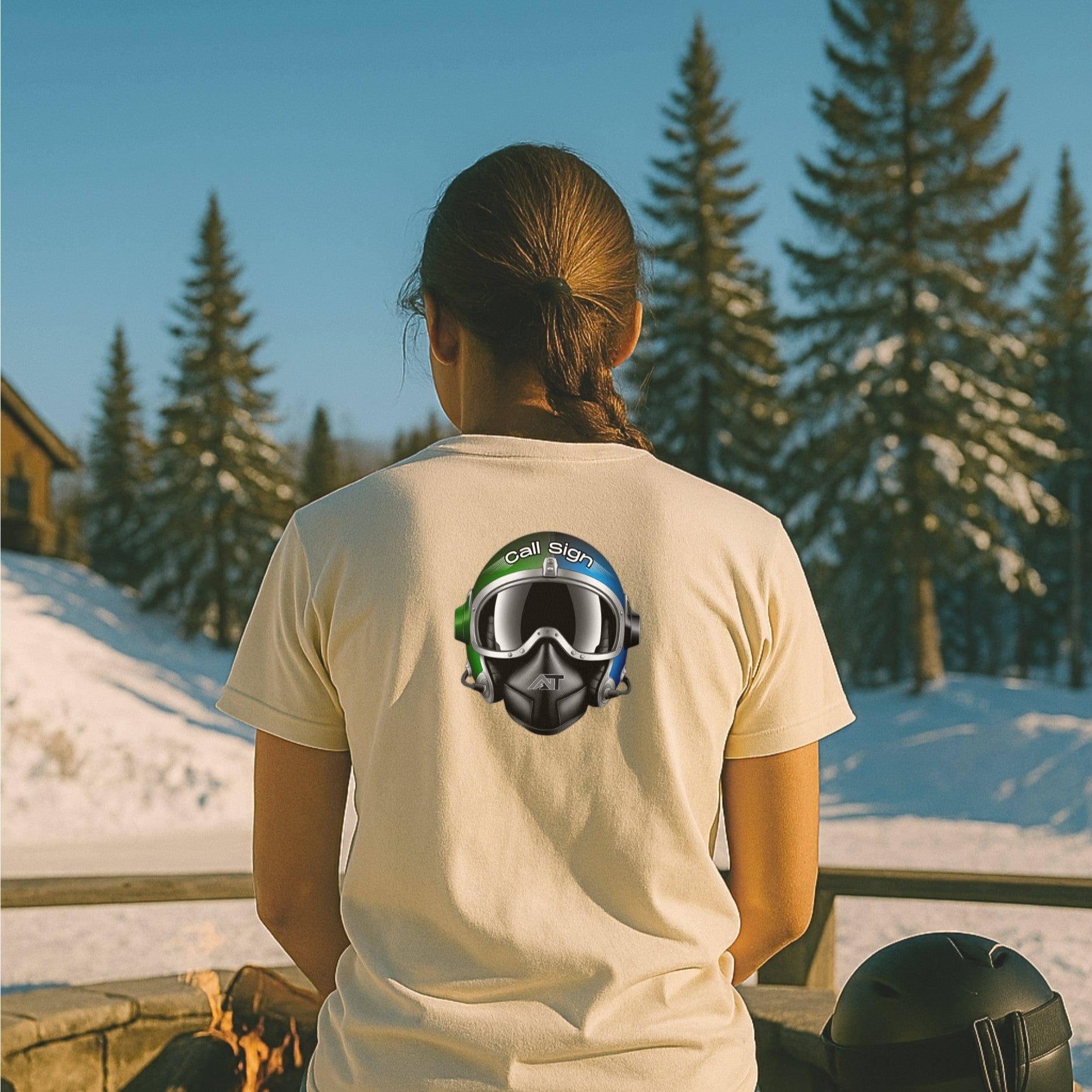 Call Sign tee from nice turns in natural, featuring the flight helmet graphic, worn at a ski lodge patio with snow and pines in the background.