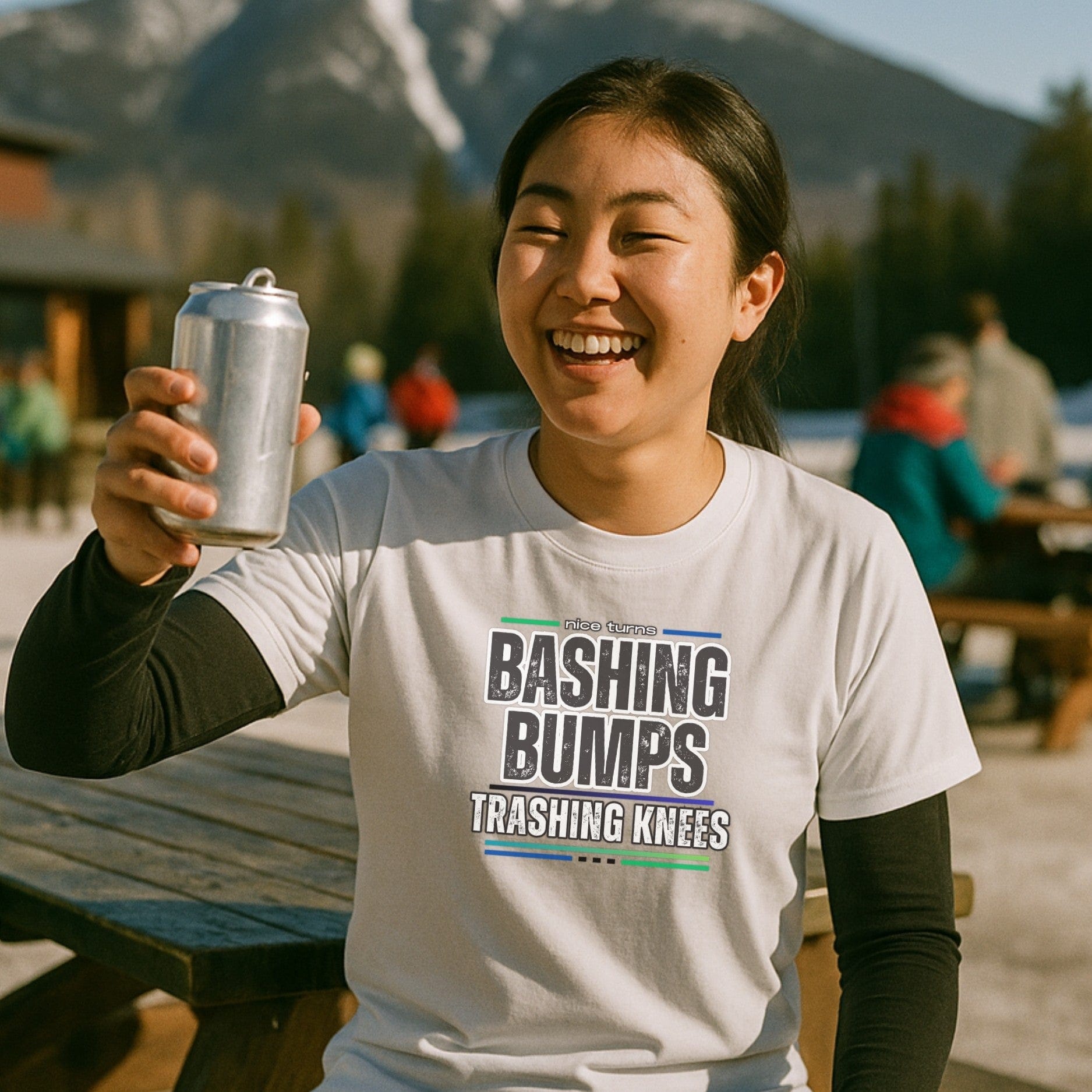 Smiling skier in a “Bashing Bumps Trashing Knees” nice turns tee, raising a can at a sunny après scene with snowy peaks behind.