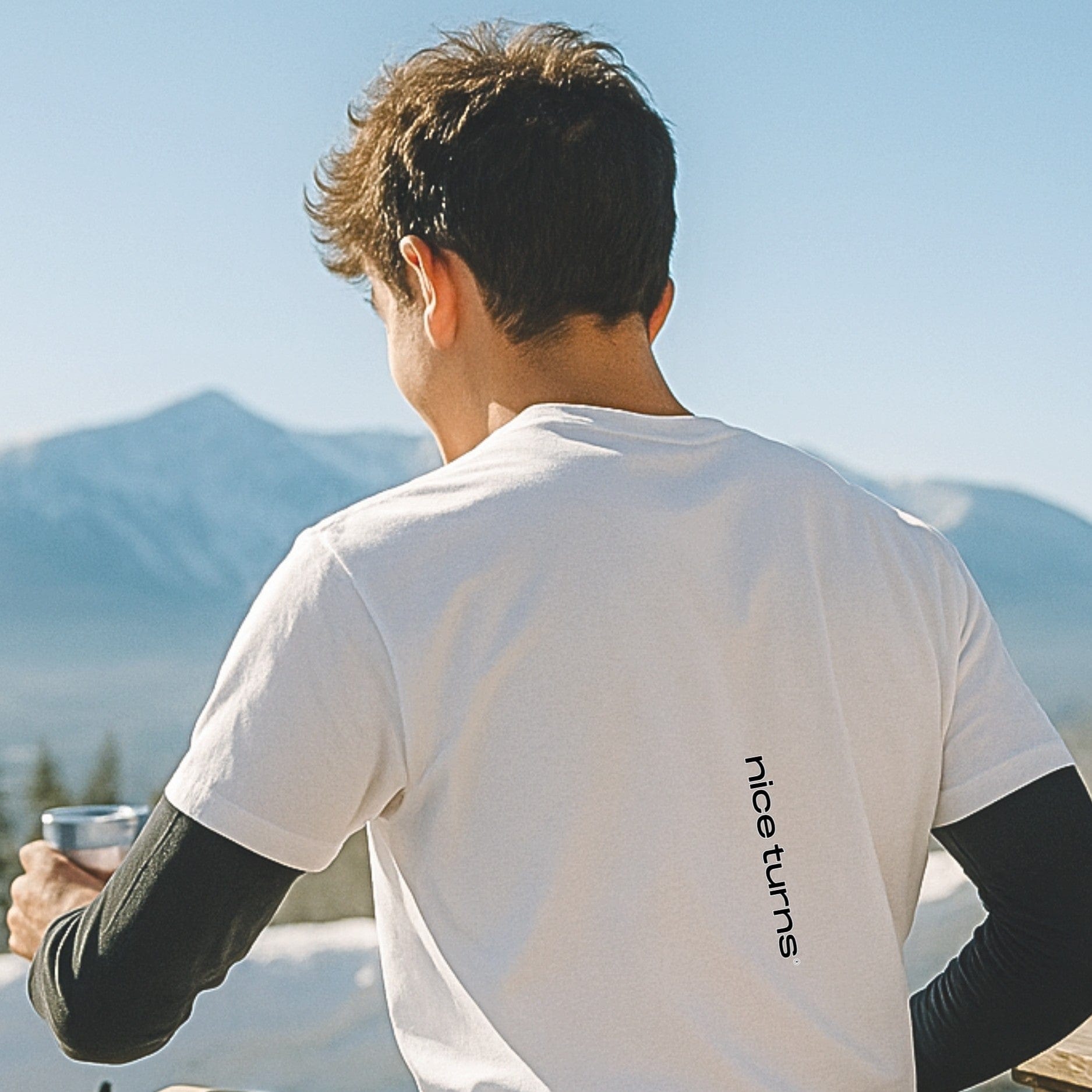 White t-shirt with vertical nice turns logo on the lower back, worn outdoors on a sunny ski deck with mountains in the background.