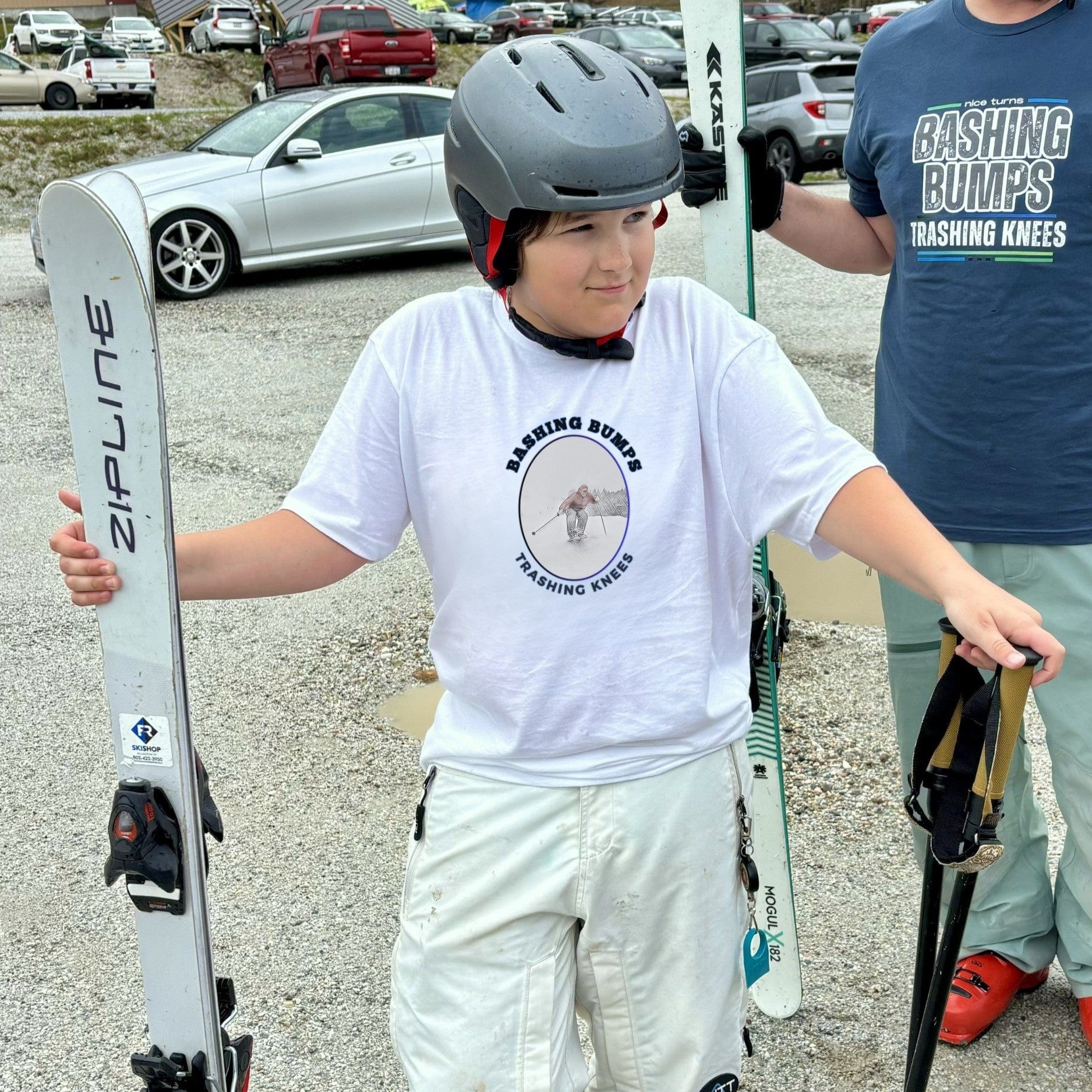 Youth skier wearing a white Bashing Bumps Trashing Knees tee in a spring ski parking lot, holding poles and Zipline mogul skis.