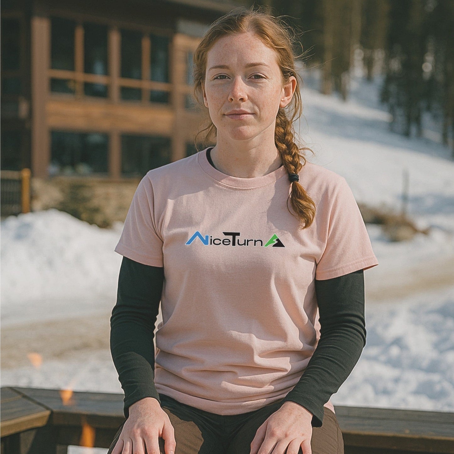 Light pink short sleeve T-shirt with the nice turns logo, worn by a skier relaxing outside a lodge on a sunny day at the mountain.