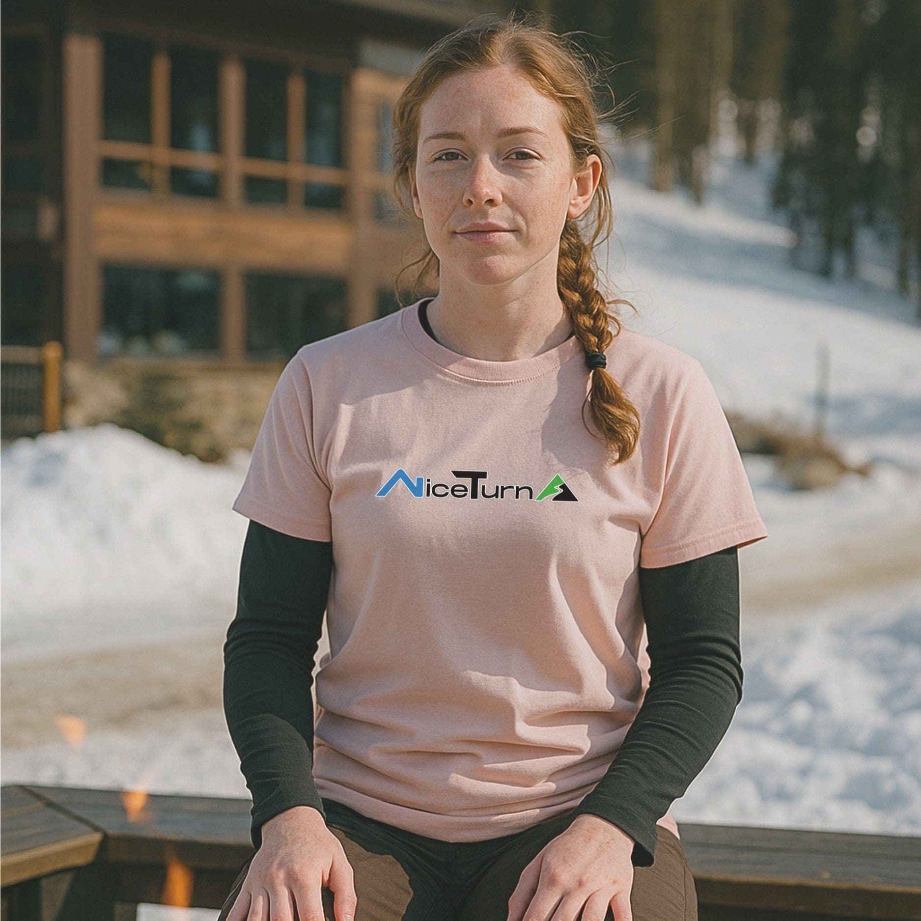 Light pink short sleeve T-shirt with the nice turns logo, worn by a skier relaxing outside a lodge on a sunny day at the mountain.