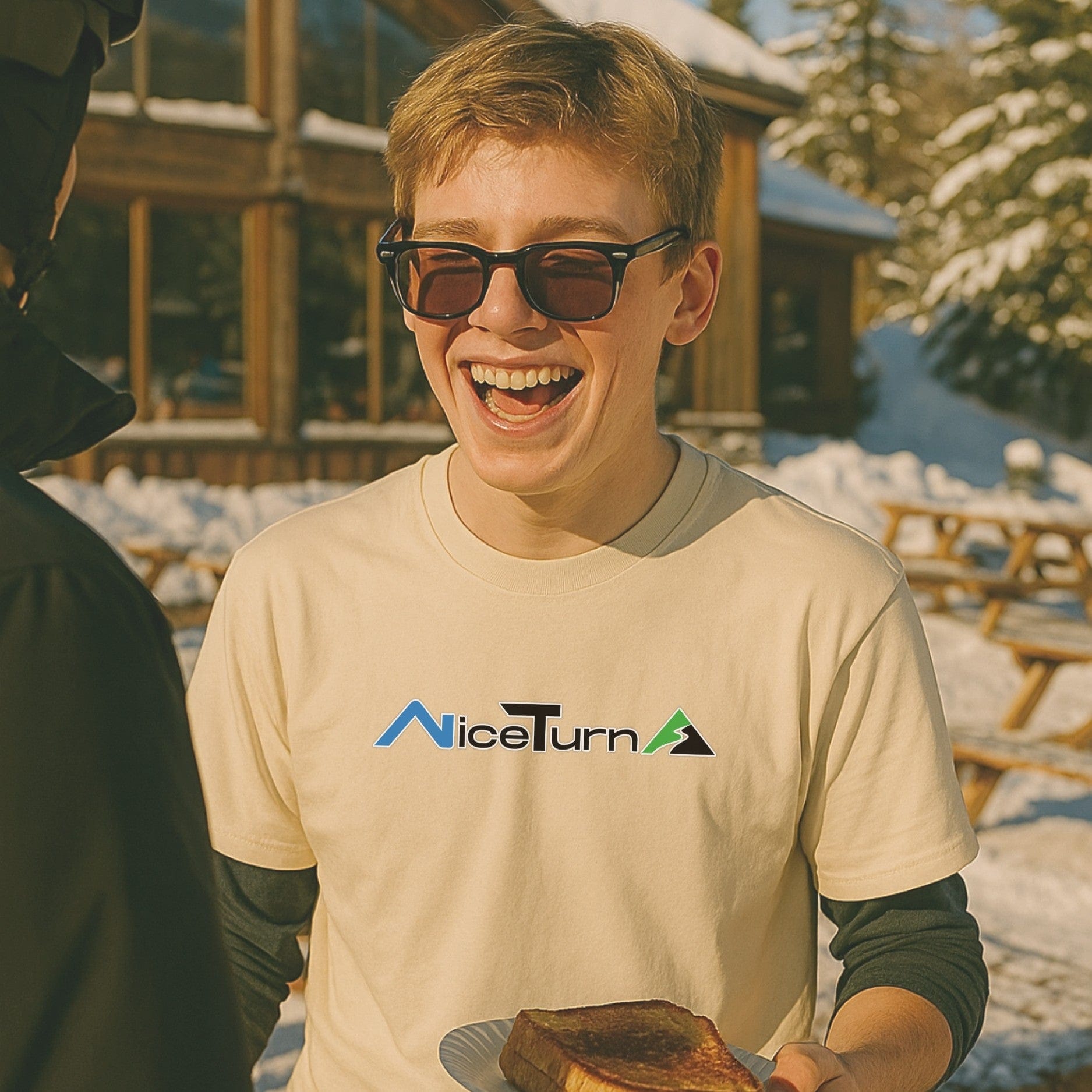 Natural short sleeve nice turns logo tee worn during a sunny après moment outside a lodge, skier laughing with grilled cheese in hand.