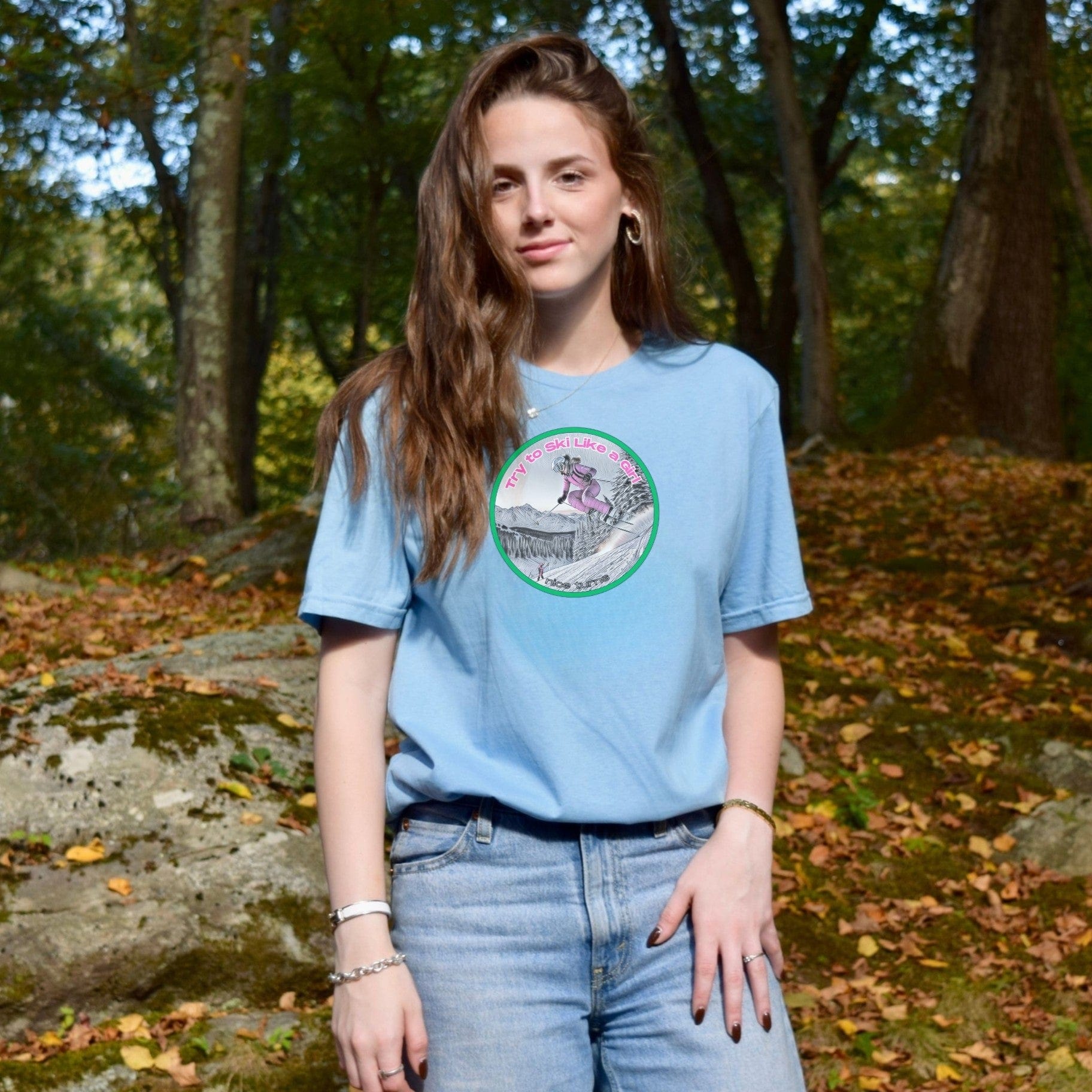 Light blue t-shirt with "Try to Ski Like a Girl" graphic, worn by a woman standing in a wooded area with early fall leaves.