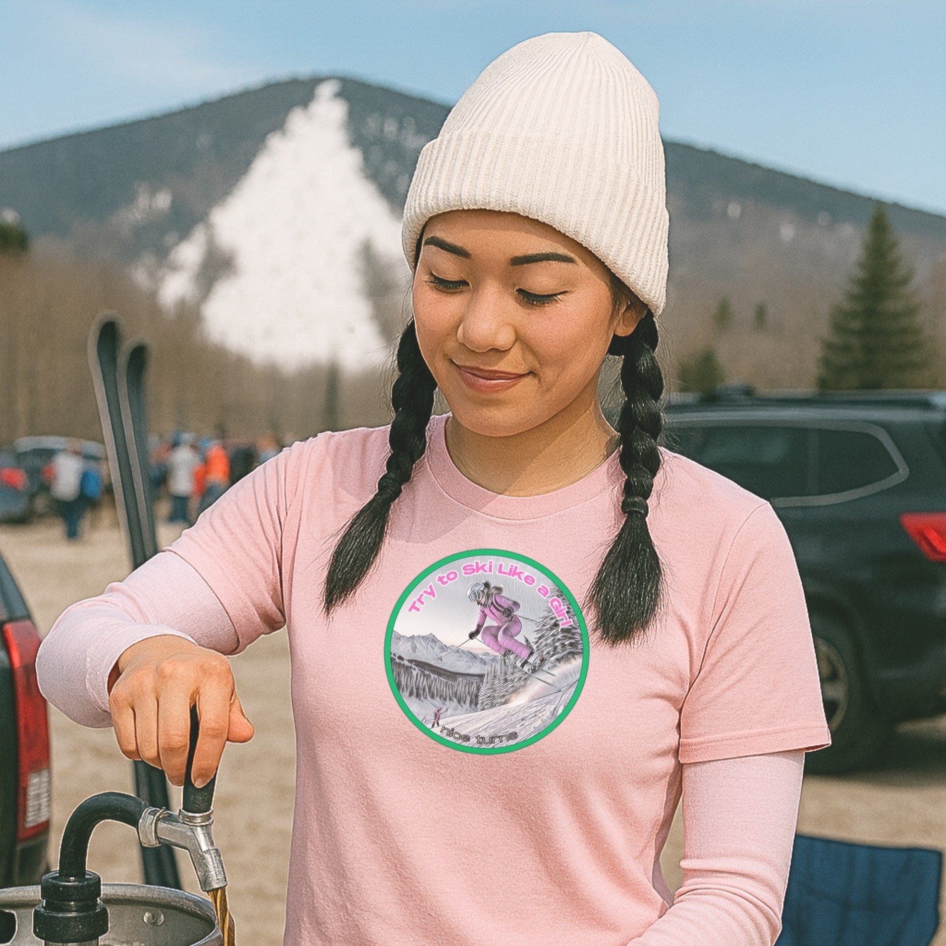 Pink t-shirt with "Try to Ski Like a Girl" skier graphic, worn by a woman pouring drinks in a ski resort parking lot with snow in the background.