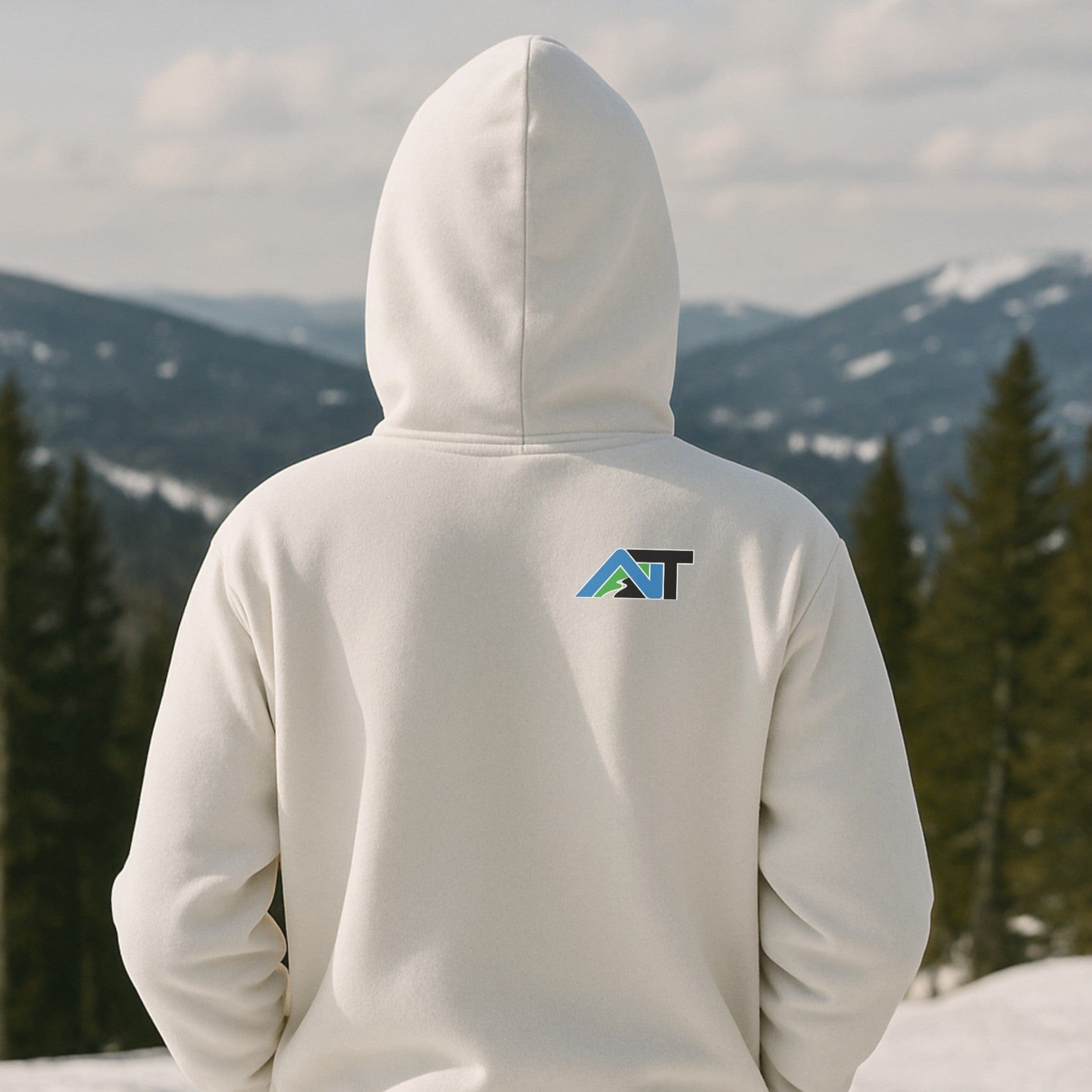 White hoodie with mountain-style AT logo on the back, worn on a snowy ridgeline with pine trees and distant alpine views.
