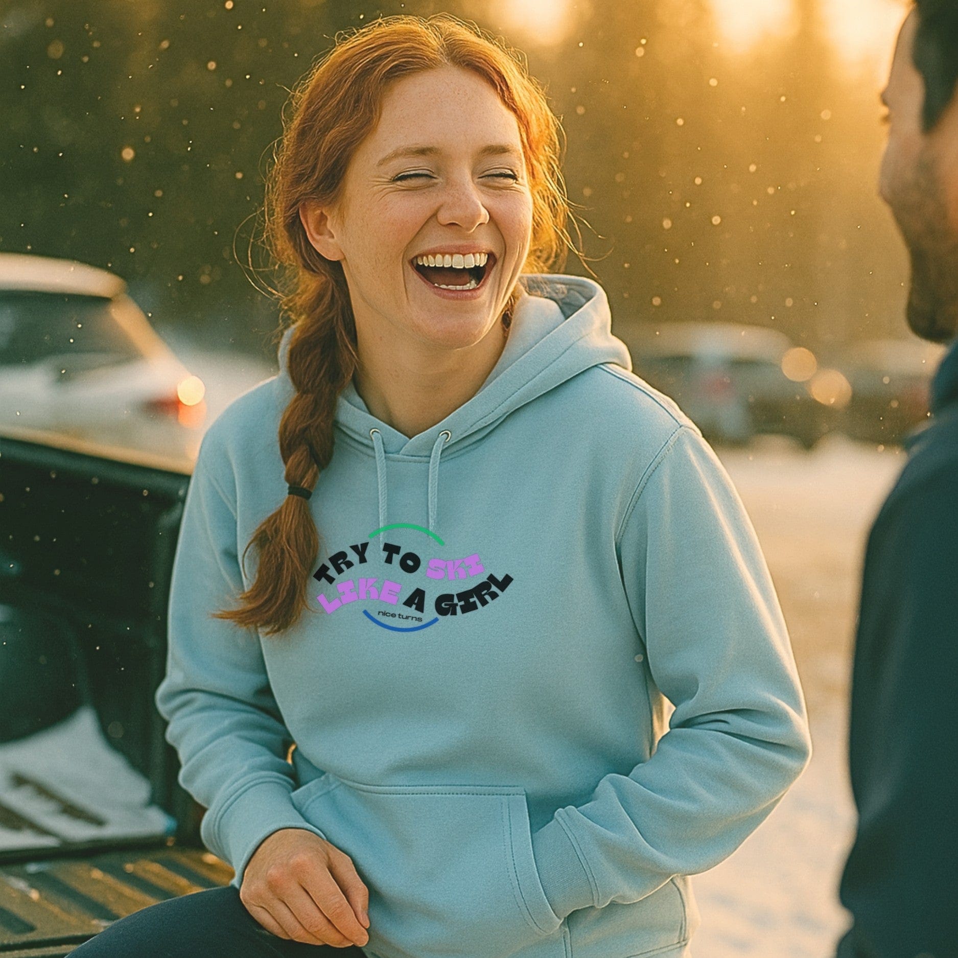Light blue hoodie with “try to ski like a girl” arc design worn at sunset in a ski parking lot. Laughter, snowflakes, and good vibes.