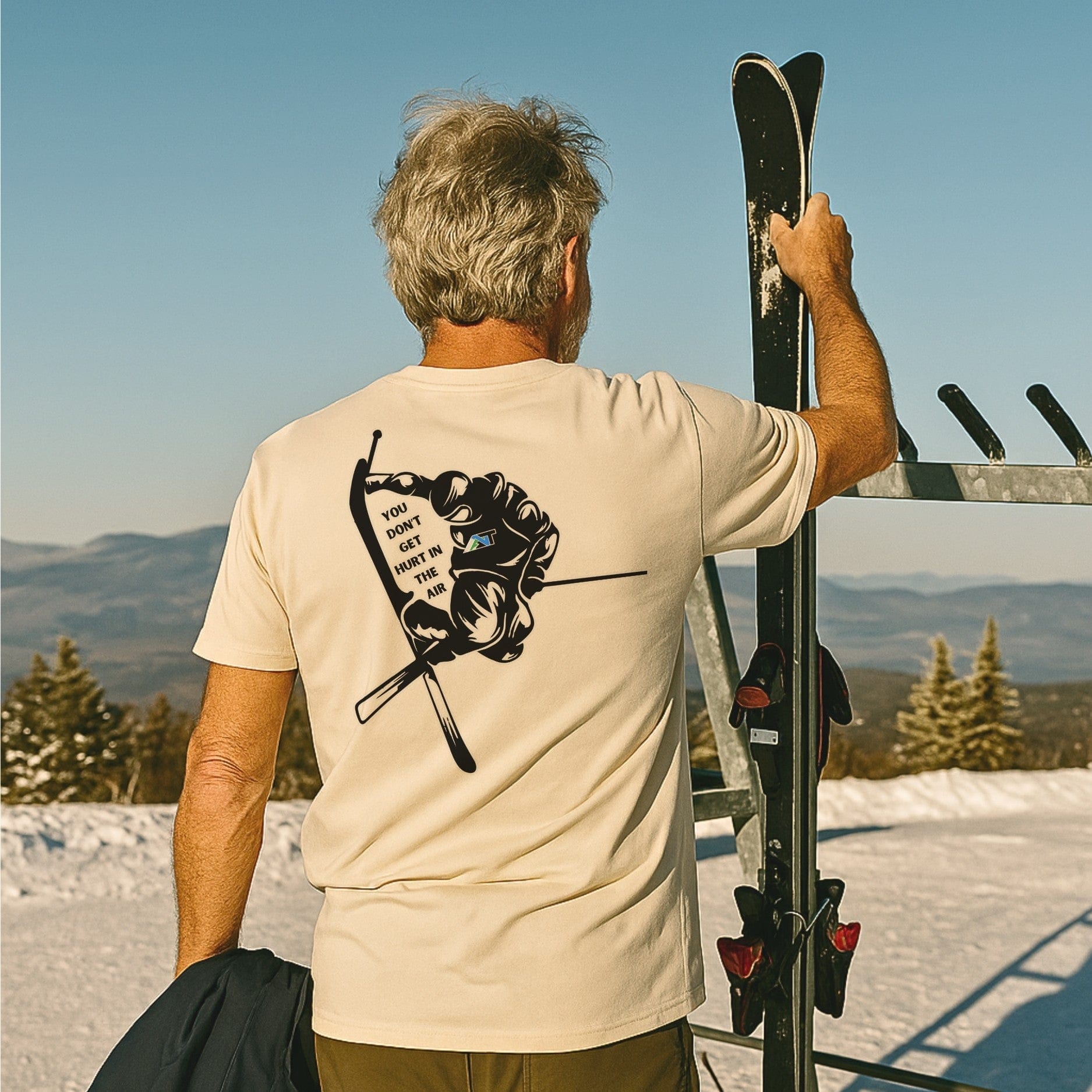 older skier placing skis at summit rack while wearing cream ski T-shirt with graphic back print