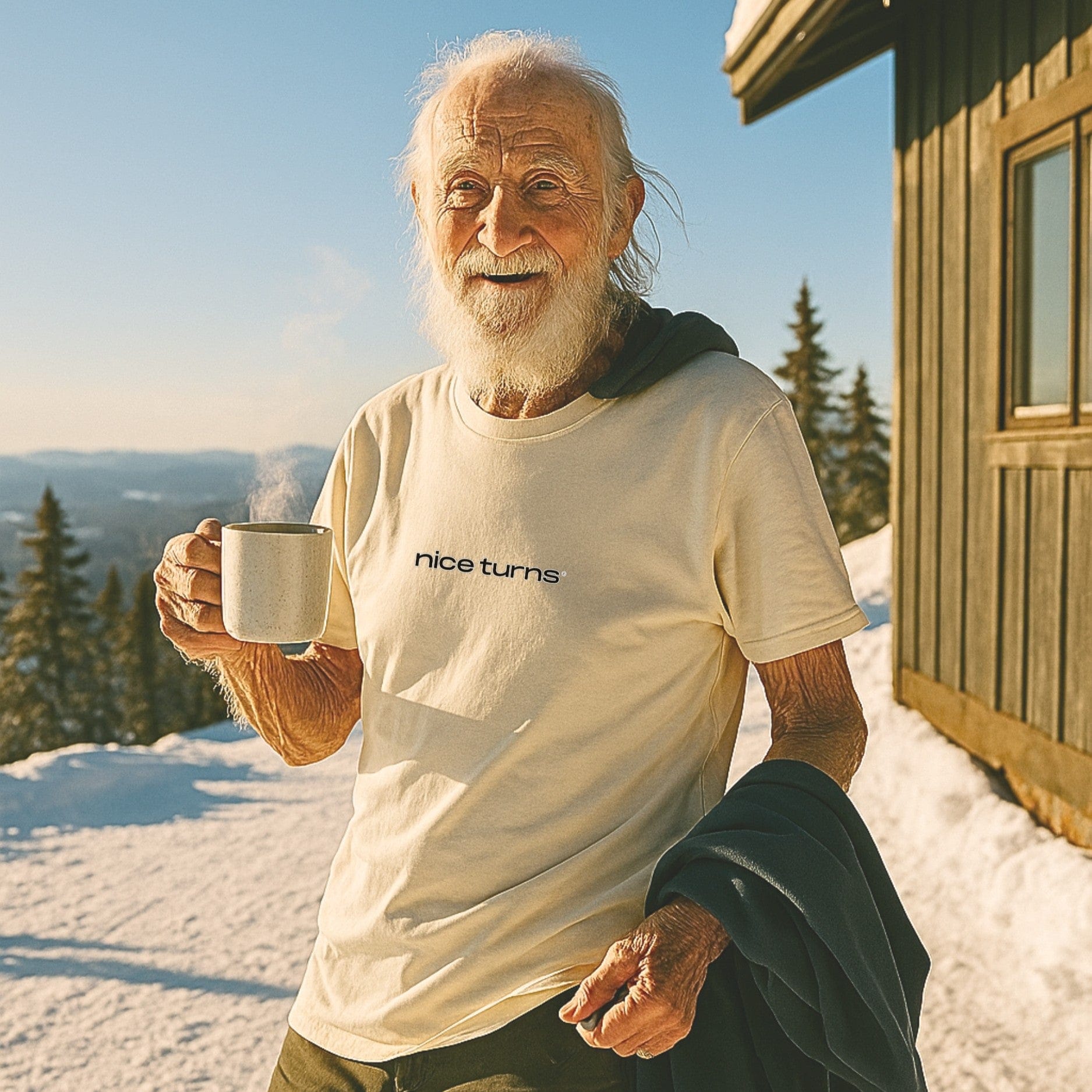 older skier wearing cream nice turns T-shirt holding coffee on sunny ski deck in spring snow